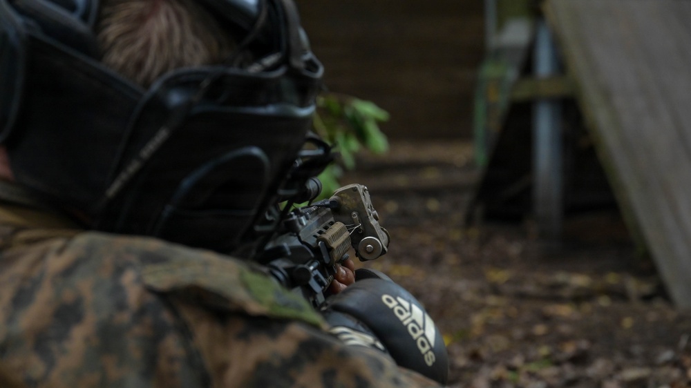 A person wearing camouflage clothing and a black protective mask is lying prone on the ground, aiming a replica firearm equipped with tactical accessories. The individual is wearing Adidas branded clothing visible on their arm, suggesting this is a recreational airsoft or paintball activity rather than a military operation. The setting appears to be an outdoor woodland environment.