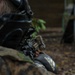 A person wearing camouflage clothing and a black protective mask is lying prone on the ground, aiming a replica firearm equipped with tactical accessories. The individual is wearing Adidas branded clothing visible on their arm, suggesting this is a recreational airsoft or paintball activity rather than a military operation. The setting appears to be an outdoor woodland environment.