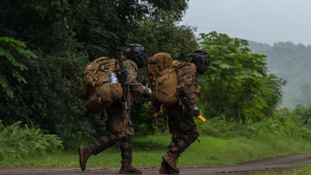 Two heavily equipped military personnel are running along a paved road in a lush, tropical or subtropical environment under overcast, misty conditions. Both soldiers are wearing full combat gear including large rucksacks, tactical helmets with face protection, and camouflage uniforms. One soldier is visibly carrying a rifle slung across their back.