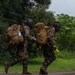 Two heavily equipped military personnel are running along a paved road in a lush, tropical or subtropical environment under overcast, misty conditions. Both soldiers are wearing full combat gear including large rucksacks, tactical helmets with face protection, and camouflage uniforms. One soldier is visibly carrying a rifle slung across their back.