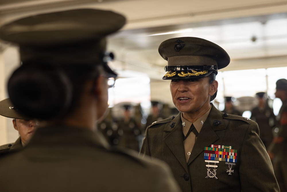 A U.S. Marine Corps officer in Service 'C' uniform with significant medal ribbons and rank insignia is engaged in conversation with another uniformed Marine. The officer facing the camera wears a peaked cap with gold scrambled eggs indicating senior officer rank, along with multiple service ribbons and decorations. The setting appears to be an indoor military facility with additional uniformed personnel visible in the background.