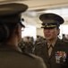 A U.S. Marine Corps officer in Service 'C' uniform with significant medal ribbons and rank insignia is engaged in conversation with another uniformed Marine. The officer facing the camera wears a peaked cap with gold scrambled eggs indicating senior officer rank, along with multiple service ribbons and decorations. The setting appears to be an indoor military facility with additional uniformed personnel visible in the background.