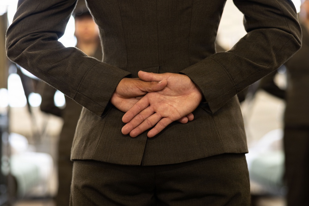 The image shows the torso and back of a person wearing a dark olive/charcoal suit, with their hands clasped together behind their back. The focus is on the hands and suit jacket, with a blurred background suggesting an indoor environment with other people present. The composition is a close-up rear view, emphasizing posture and body language.