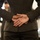 Alpha Company Battalion Commander’s Inspection The image shows the torso and back of a person wearing a dark olive/charcoal suit, with their hands clasped together behind their back. The focus is on the hands and suit jacket, with a blurred background suggesting an indoor environment with other people present. The composition is a close-up rear view, emphasizing posture and body language.