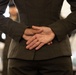The image shows the torso and back of a person wearing a dark olive/charcoal suit, with their hands clasped together behind their back. The focus is on the hands and suit jacket, with a blurred background suggesting an indoor environment with other people present. The composition is a close-up rear view, emphasizing posture and body language.