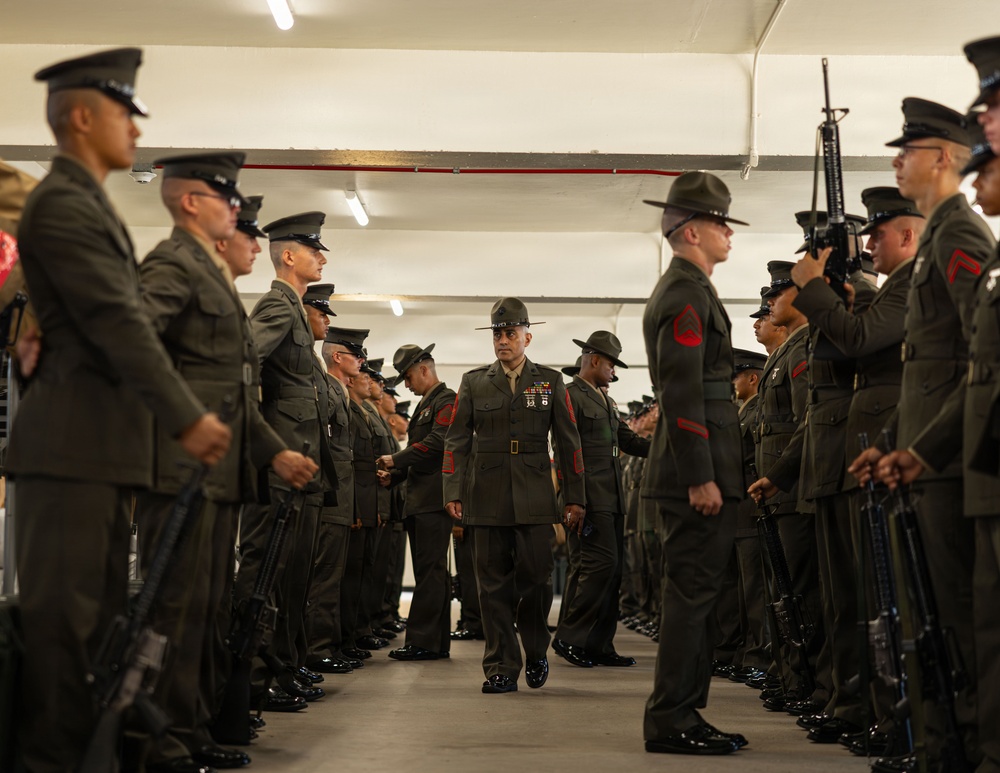 A senior U.S. Marine Corps officer or senior NCO walks through a corridor formed by two rows of Marines standing at attention in an indoor facility, likely a parking structure or covered drill deck. The central figure appears to be conducting an inspection or ceremonial passage, wearing dress uniform with visible medals and ribbons. Marines on both sides are in service uniforms, some holding M16/M4-style rifles.