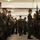 Alpha Company Battalion Commander’s Inspection A senior U.S. Marine Corps officer or senior NCO walks through a corridor formed by two rows of Marines standing at attention in an indoor facility, likely a parking structure or covered drill deck. The central figure appears to be conducting an inspection or ceremonial passage, wearing dress uniform with visible medals and ribbons. Marines on both sides are in service uniforms, some holding M16/M4-style rifles.