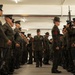 A senior U.S. Marine Corps officer or senior NCO walks through a corridor formed by two rows of Marines standing at attention in an indoor facility, likely a parking structure or covered drill deck. The central figure appears to be conducting an inspection or ceremonial passage, wearing dress uniform with visible medals and ribbons. Marines on both sides are in service uniforms, some holding M16/M4-style rifles.