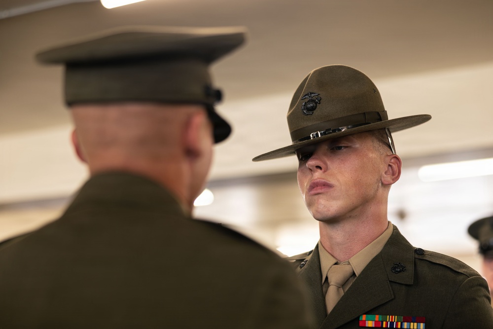 Two U.S. Marine Corps personnel are shown in a formal indoor setting, facing each other. The Marine in focus is wearing a Drill Instructor campaign cover (campaign hat) with the Eagle, Globe, and Anchor emblem, along with service ribbons on his dress uniform. The scene appears to depict a formal inspection or ceremony.