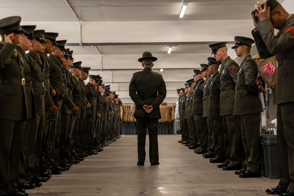 A military drill instructor or senior non-commissioned officer stands with hands clasped behind their back in the center of a formation of uniformed personnel lined up in two rows facing each other inside what appears to be a barracks or equipment room. The personnel are wearing olive drab dress uniforms with garrison caps, consistent with U.S. Marine Corps service uniforms. The scene appears to depict a formal inspection or uniform review.