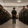 Alpha Company Battalion Commander’s Inspection A military drill instructor or senior non-commissioned officer stands with hands clasped behind their back in the center of a formation of uniformed personnel lined up in two rows facing each other inside what appears to be a barracks or equipment room. The personnel are wearing olive drab dress uniforms with garrison caps, consistent with U.S. Marine Corps service uniforms. The scene appears to depict a formal inspection or uniform review.