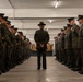 A military drill instructor or senior non-commissioned officer stands with hands clasped behind their back in the center of a formation of uniformed personnel lined up in two rows facing each other inside what appears to be a barracks or equipment room. The personnel are wearing olive drab dress uniforms with garrison caps, consistent with U.S. Marine Corps service uniforms. The scene appears to depict a formal inspection or uniform review.