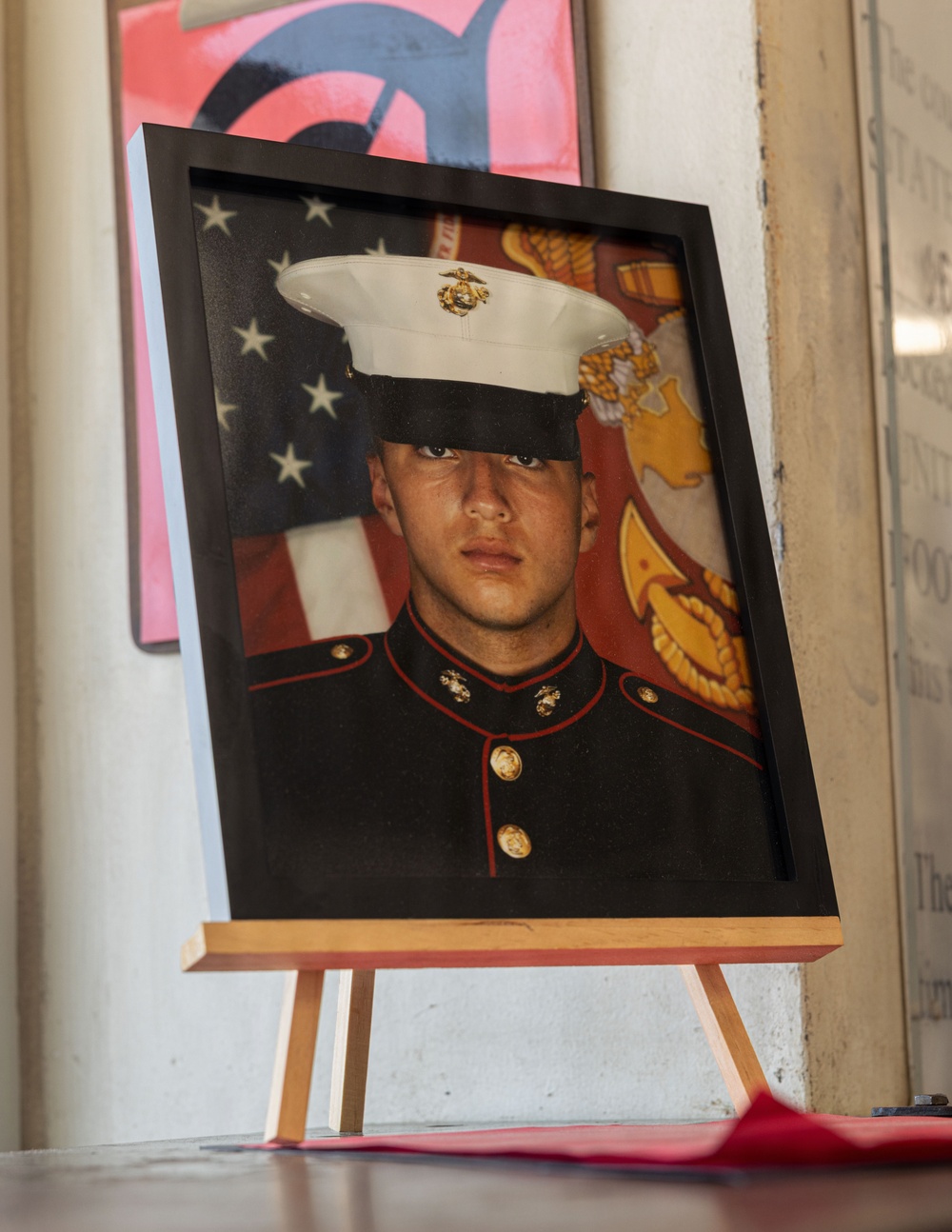 A framed portrait photograph of a young United States Marine in dress blues uniform is displayed on a wooden easel. The formal military portrait shows the subject wearing the distinctive white cover (hat) with the Eagle, Globe, and Anchor emblem, set against an American flag and Marine Corps emblem background. The display setting suggests this may be a memorial or commemorative presentation.