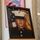 Alpha Company Battalion Commander’s Inspection A framed portrait photograph of a young United States Marine in dress blues uniform is displayed on a wooden easel. The formal military portrait shows the subject wearing the distinctive white cover (hat) with the Eagle, Globe, and Anchor emblem, set against an American flag and Marine Corps emblem background. The display setting suggests this may be a memorial or commemorative presentation.