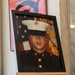 A framed portrait photograph of a young United States Marine in dress blues uniform is displayed on a wooden easel. The formal military portrait shows the subject wearing the distinctive white cover (hat) with the Eagle, Globe, and Anchor emblem, set against an American flag and Marine Corps emblem background. The display setting suggests this may be a memorial or commemorative presentation.