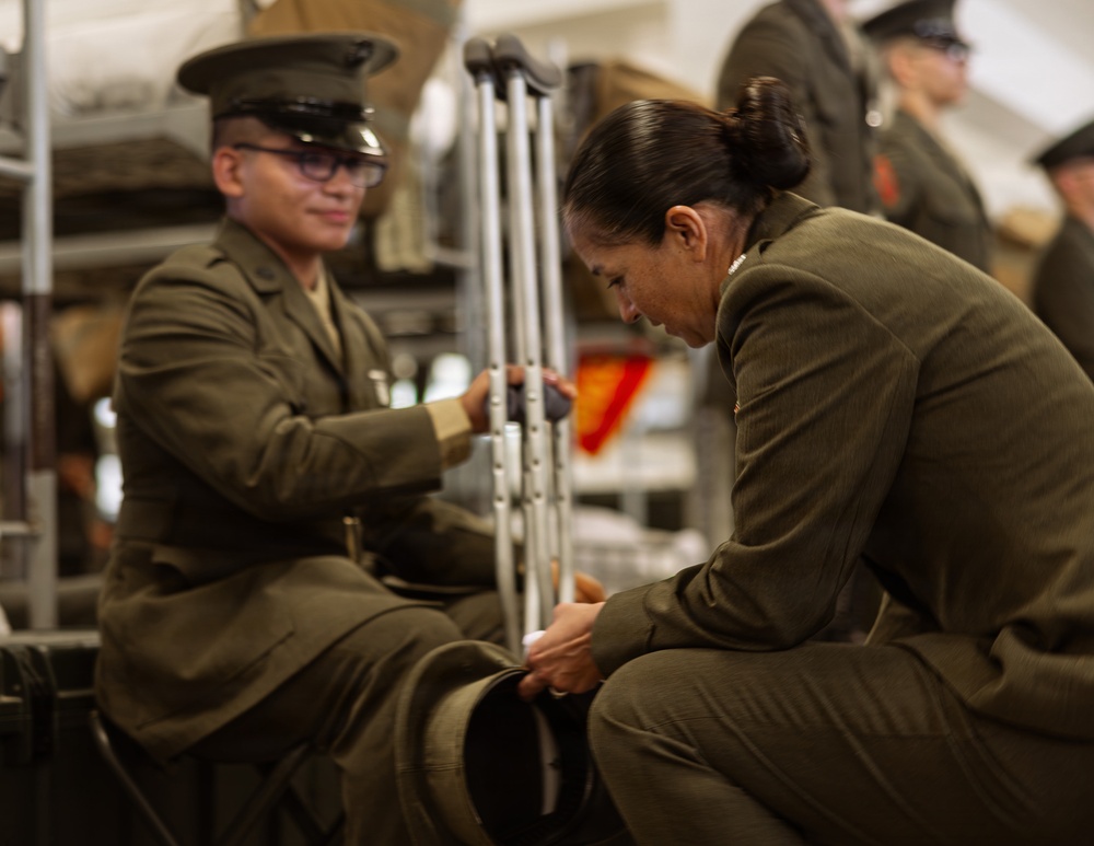 Two military personnel in olive/green dress uniforms are engaged in what appears to be a uniform inspection or assistance scenario. One service member is seated and using crutches, suggesting an injury, while the other is crouched down attending to the seated individual's lower leg or boot area. Additional uniformed personnel are visible in the blurred background.