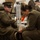 Alpha Company Battalion Commander’s Inspection Two military personnel in olive/green dress uniforms are engaged in what appears to be a uniform inspection or assistance scenario. One service member is seated and using crutches, suggesting an injury, while the other is crouched down attending to the seated individual's lower leg or boot area. Additional uniformed personnel are visible in the blurred background.