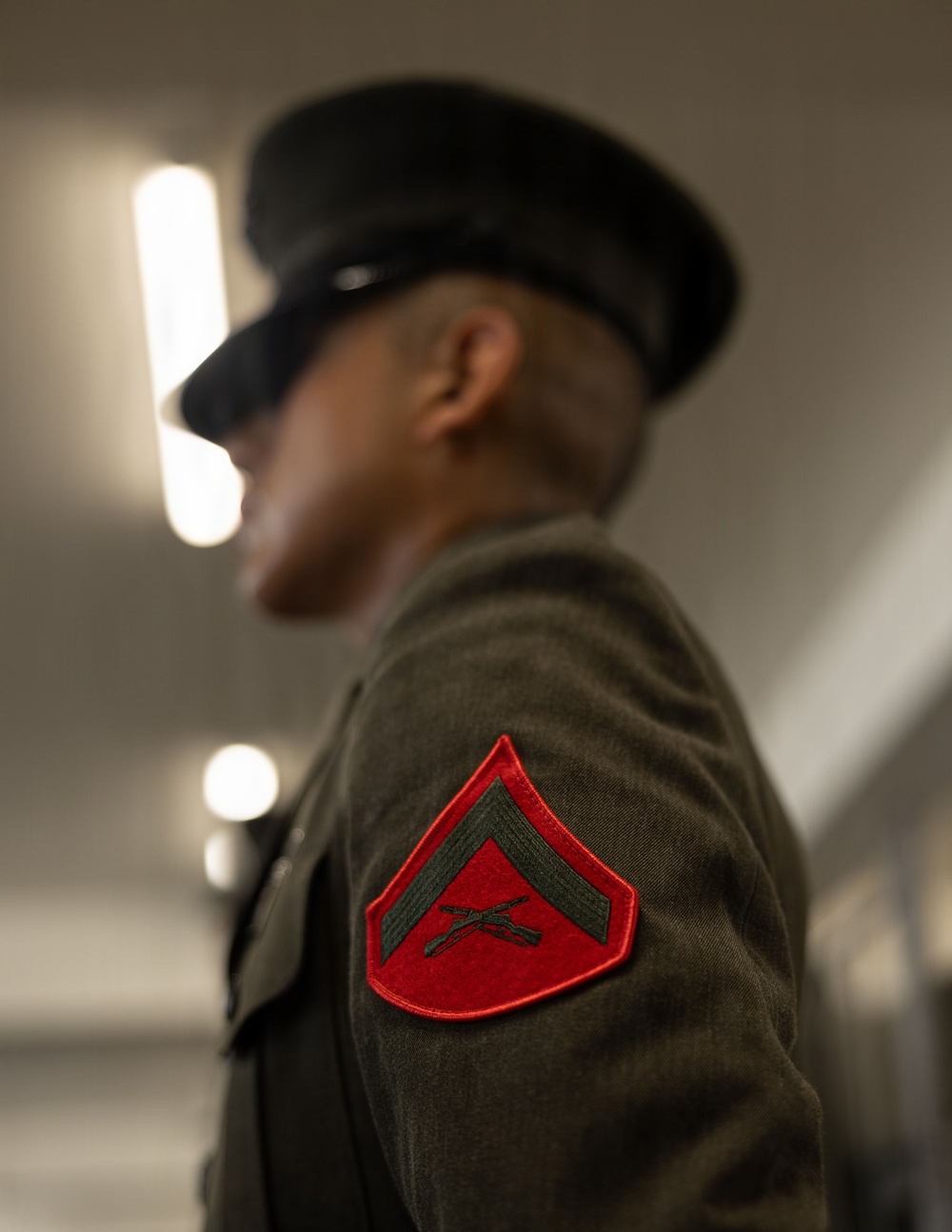 A close-up photograph focusing on the sleeve insignia of a United States Marine Corps dress uniform. The patch is a red chevron with crossed rifles, indicating the rank of Lance Corporal (E-3). The subject's face is intentionally blurred in the background, with the rank patch as the sharp focal point.