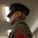 A close-up photograph focusing on the sleeve insignia of a United States Marine Corps dress uniform. The patch is a red chevron with crossed rifles, indicating the rank of Lance Corporal (E-3). The subject's face is intentionally blurred in the background, with the rank patch as the sharp focal point.