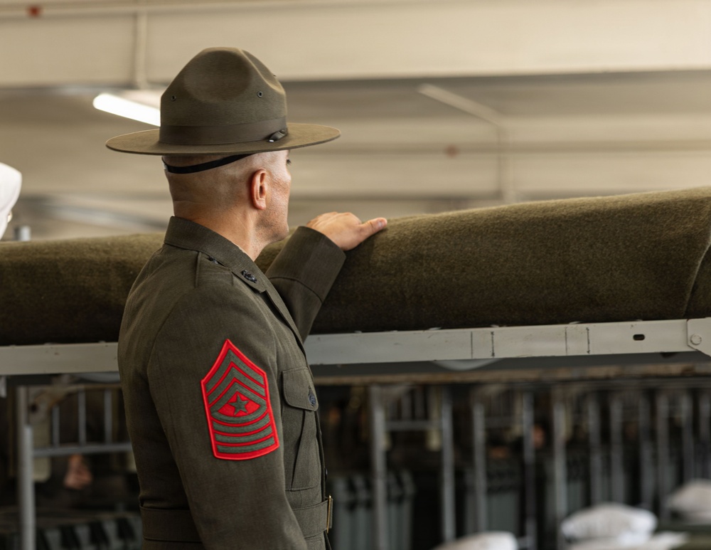 A U.S. Marine Corps Sergeant Major is shown from behind, wearing the green service uniform and a drill instructor's campaign cover (campaign hat). He is inspecting or pointing at what appears to be rolled green wool blankets or fabric stored on metal racks. The setting appears to be a military supply or storage facility.