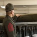 A U.S. Marine Corps Sergeant Major is shown from behind, wearing the green service uniform and a drill instructor's campaign cover (campaign hat). He is inspecting or pointing at what appears to be rolled green wool blankets or fabric stored on metal racks. The setting appears to be a military supply or storage facility.