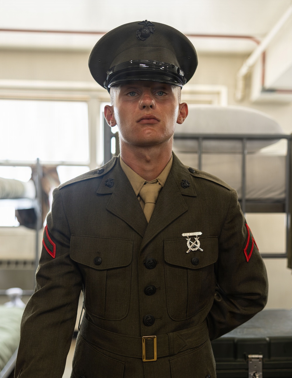 A young male United States Marine is photographed in Service Alpha (Dress Green) uniform inside what appears to be a military barracks. He is wearing the rank insignia of a Private First Class (PFC) on both sleeves and a Rifle Expert badge on his chest. The image is a sharp, well-lit portrait-style photograph taken in an indoor military setting.