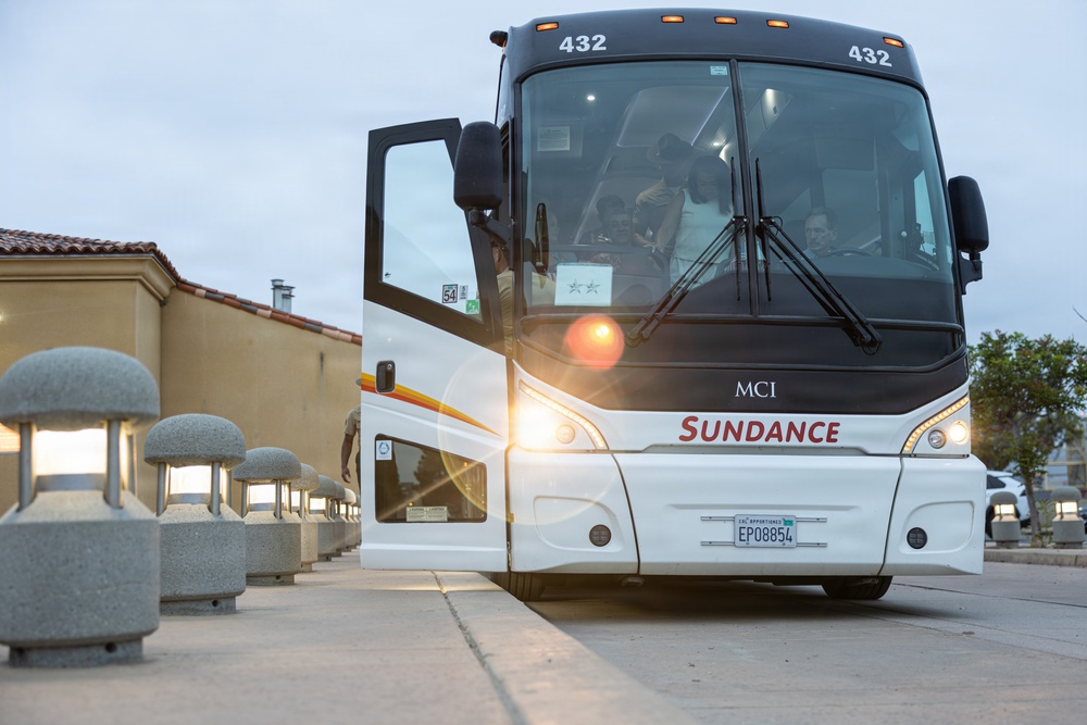 A white MCI motorcoach operated by Sundance, numbered 432, is approaching the camera on a paved driveway or terminal area. The bus has its headlights on, creating a lens flare effect, and passengers are visible through the windshield. The setting appears to be a hotel or transit facility at dusk or dawn.
