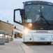 A white MCI motorcoach operated by Sundance, numbered 432, is approaching the camera on a paved driveway or terminal area. The bus has its headlights on, creating a lens flare effect, and passengers are visible through the windshield. The setting appears to be a hotel or transit facility at dusk or dawn.