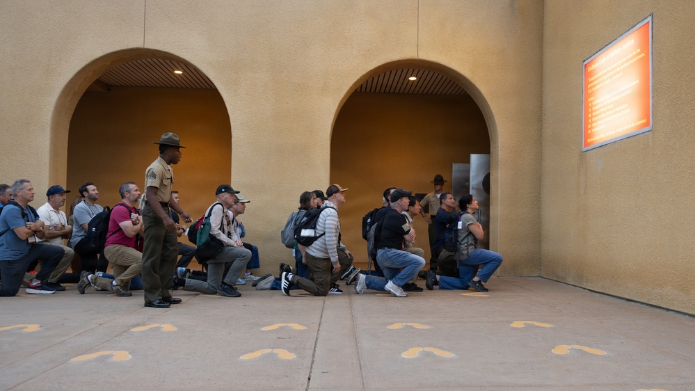 A group of civilians is kneeling on a concrete outdoor area while a uniformed military drill instructor stands and addresses them. Yellow footprint markings are painted on the ground, which is a hallmark of Marine Corps recruit training orientation areas. A second uniformed figure is visible in the background near an archway, and an orange informational sign is mounted on the wall to the right.