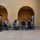 A group of civilians is kneeling on a concrete outdoor area while a uniformed military drill instructor stands and addresses them. Yellow footprint markings are painted on the ground, which is a hallmark of Marine Corps recruit training orientation areas. A second uniformed figure is visible in the background near an archway, and an orange informational sign is mounted on the wall to the right.