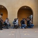A group of civilians is kneeling on a concrete outdoor area while a uniformed military drill instructor stands and addresses them. Yellow footprint markings are painted on the ground, which is a hallmark of Marine Corps recruit training orientation areas. A second uniformed figure is visible in the background near an archway, and an orange informational sign is mounted on the wall to the right.
