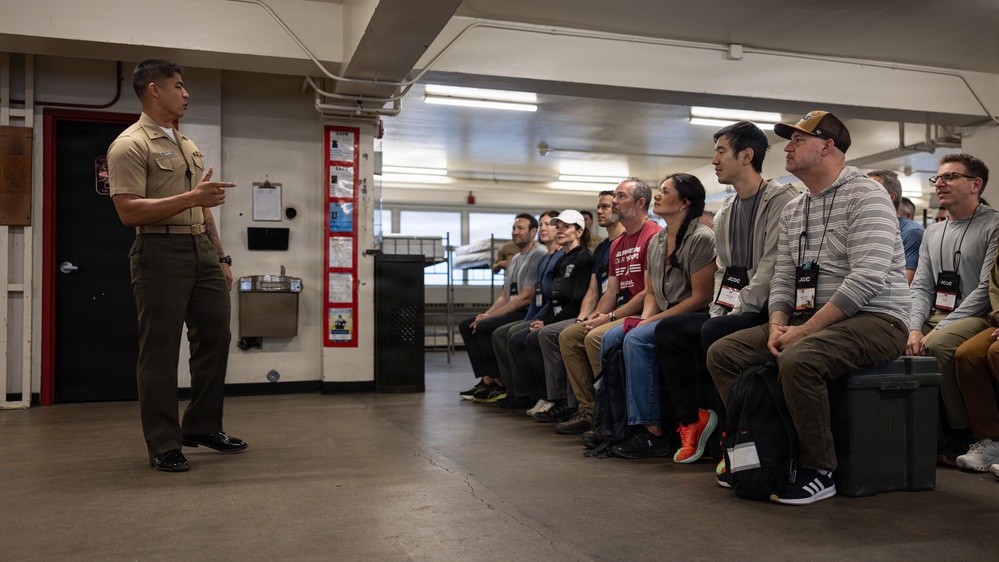 A U.S. Marine in service uniform (tan/khaki) is addressing a group of approximately 12-15 civilians seated along a wall in what appears to be a military barracks or training facility. The civilians are wearing lanyards with badges, suggesting they are visitors on an organized tour or orientation program. The Marine appears to be giving a briefing or presentation to the group.