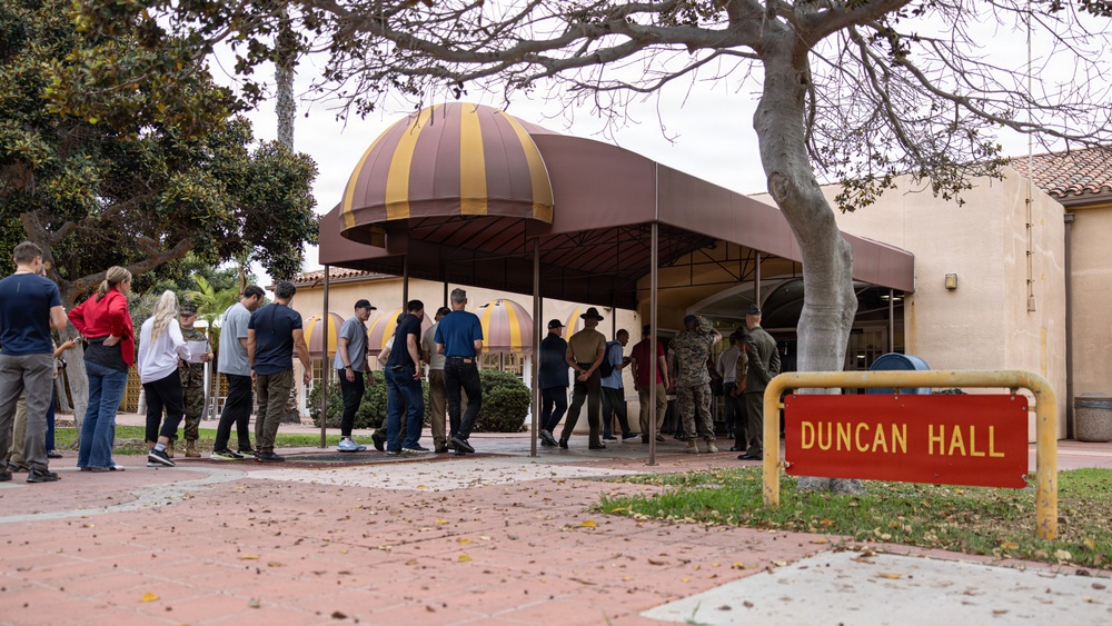 A group of civilians and uniformed military personnel are gathered outside Duncan Hall, a building with a distinctive dome-shaped awning in maroon and gold stripes. The scene appears to depict a military processing or orientation event, with civilians lined up and military personnel in camouflage uniforms overseeing the activity. A red and yellow 'Duncan Hall' sign is prominently visible in the foreground.