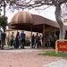 A group of civilians and uniformed military personnel are gathered outside Duncan Hall, a building with a distinctive dome-shaped awning in maroon and gold stripes. The scene appears to depict a military processing or orientation event, with civilians lined up and military personnel in camouflage uniforms overseeing the activity. A red and yellow 'Duncan Hall' sign is prominently visible in the foreground.