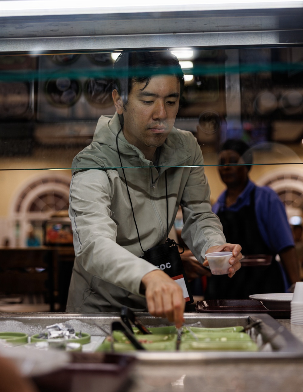 A man in a light gray zip-up jacket is leaning over a food service counter, reaching toward food items with one hand while holding a small plastic cup in the other. The scene appears to be inside a cafeteria or food service establishment, with a sneeze guard glass panel visible in the foreground. A second person wearing a dark apron and blue shirt is visible in the background.