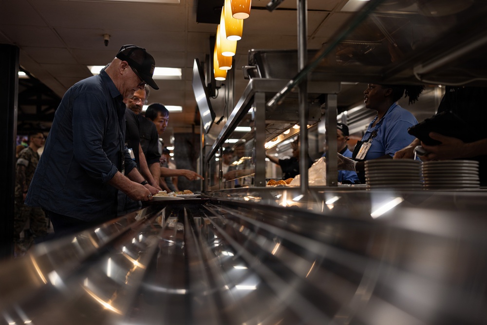 The image depicts a cafeteria or military mess hall serving line, where several individuals are being served food by staff behind a stainless steel counter. Warm overhead heat lamps illuminate the food service area, and a mix of civilians and what appear to be military personnel are present. The scene suggests an institutional dining facility, possibly on a military base.