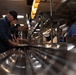 The image depicts a cafeteria or military mess hall serving line, where several individuals are being served food by staff behind a stainless steel counter. Warm overhead heat lamps illuminate the food service area, and a mix of civilians and what appear to be military personnel are present. The scene suggests an institutional dining facility, possibly on a military base.