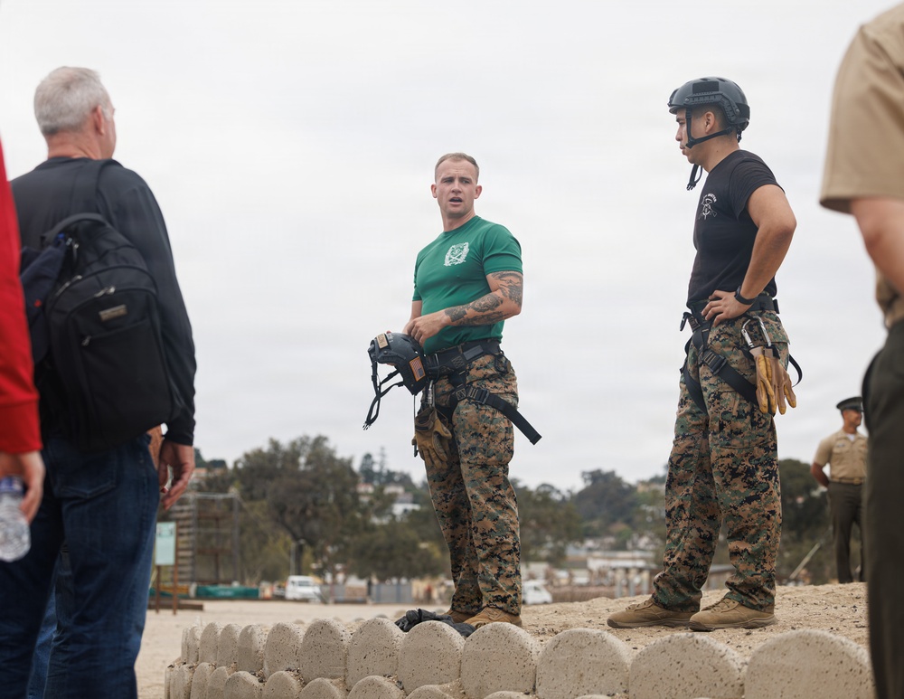 Two military personnel in MARPAT camouflage trousers stand on a low concrete barrier structure, appearing to brief or instruct a small group of civilians. One service member wears a green t-shirt and holds a helmet, while the other wears a black t-shirt and tactical helmet with a climbing harness. The setting appears to be an outdoor military training or demonstration area.