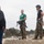 Two military personnel in MARPAT camouflage trousers stand on a low concrete barrier structure, appearing to brief or instruct a small group of civilians. One service member wears a green t-shirt and holds a helmet, while the other wears a black t-shirt and tactical helmet with a climbing harness. The setting appears to be an outdoor military training or demonstration area.