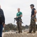 Two military personnel in MARPAT camouflage trousers stand on a low concrete barrier structure, appearing to brief or instruct a small group of civilians. One service member wears a green t-shirt and holds a helmet, while the other wears a black t-shirt and tactical helmet with a climbing harness. The setting appears to be an outdoor military training or demonstration area.