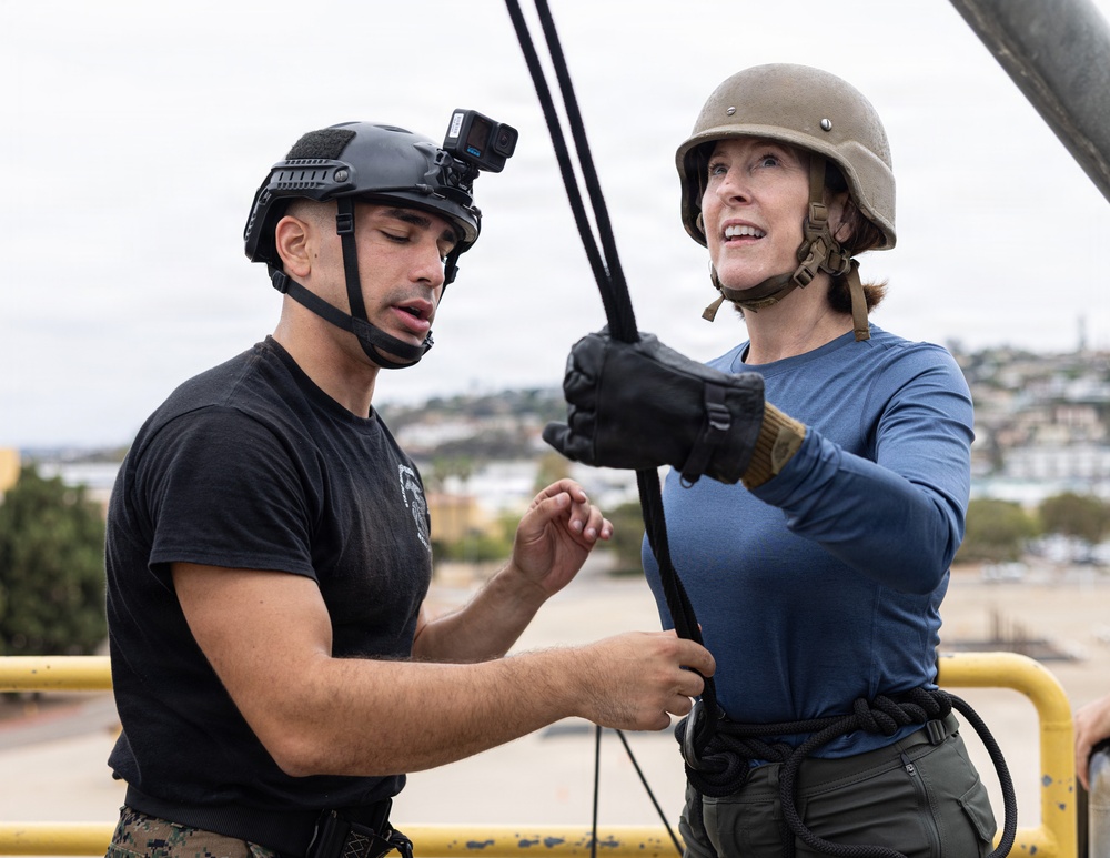 Two individuals are on an elevated platform, appearing to be engaged in a rappelling or rope descent training exercise. A person in military-style attire with a black tactical helmet mounted with a GoPro camera is assisting a woman wearing a tan helmet and blue long-sleeve shirt who is gripping a rope with gloved hands. The setting appears to be a military or training facility with a coastal urban backdrop visible in the background.