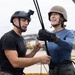Two individuals are on an elevated platform, appearing to be engaged in a rappelling or rope descent training exercise. A person in military-style attire with a black tactical helmet mounted with a GoPro camera is assisting a woman wearing a tan helmet and blue long-sleeve shirt who is gripping a rope with gloved hands. The setting appears to be a military or training facility with a coastal urban backdrop visible in the background.