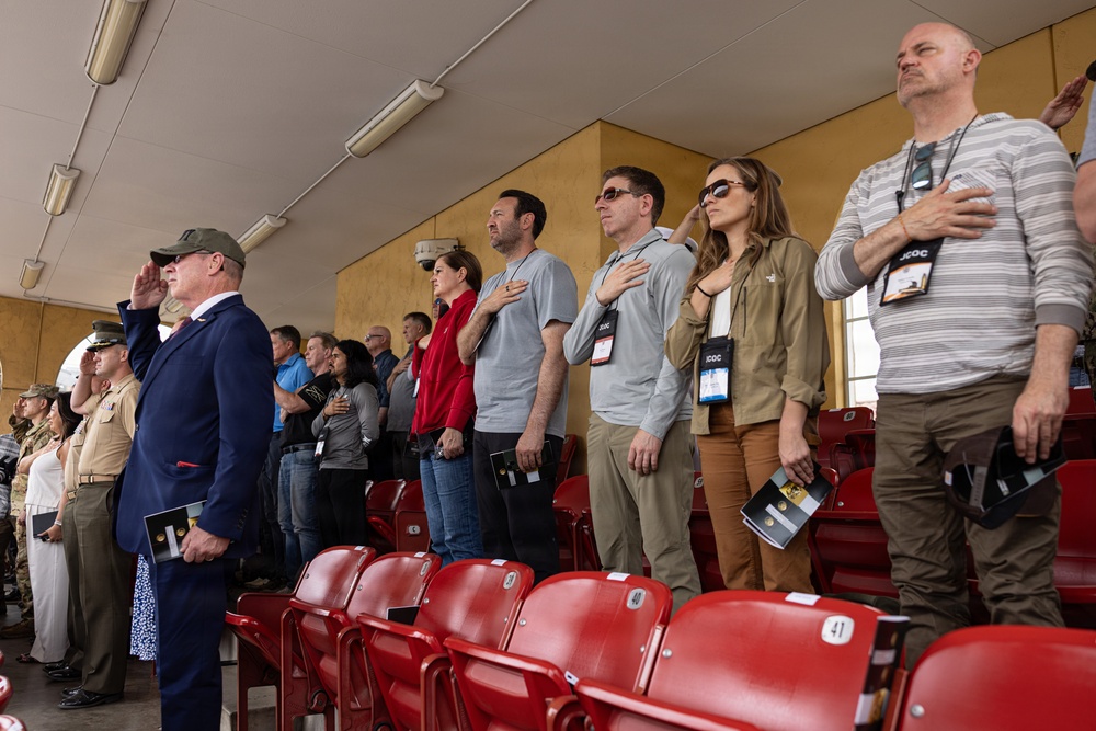 A group of people standing in stadium bleachers during what appears to be a national anthem or pledge ceremony. Several individuals have their right hands placed over their hearts, while one uniformed military officer on the left renders a salute. The setting appears to be an indoor or covered stadium with red seating.