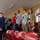 A group of people standing in stadium bleachers during what appears to be a national anthem or pledge ceremony. Several individuals have their right hands placed over their hearts, while one uniformed military officer on the left renders a salute. The setting appears to be an indoor or covered stadium with red seating.