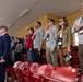 A group of people standing in stadium bleachers during what appears to be a national anthem or pledge ceremony. Several individuals have their right hands placed over their hearts, while one uniformed military officer on the left renders a salute. The setting appears to be an indoor or covered stadium with red seating.