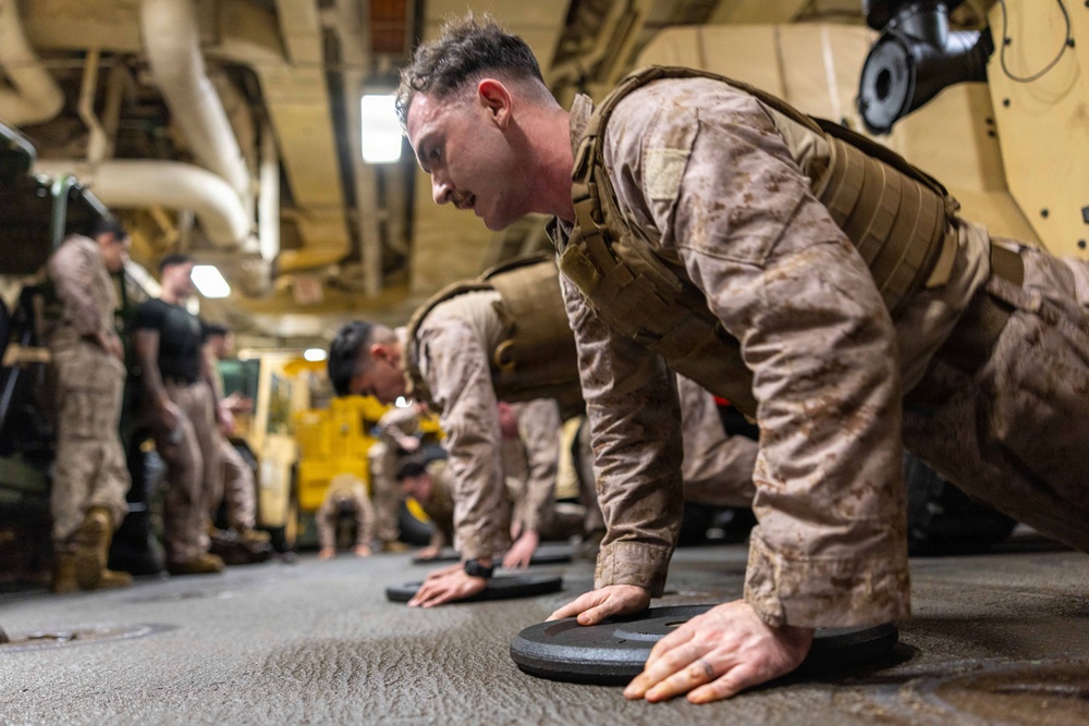 Military personnel in desert camouflage uniforms are performing push-ups on weight plates in what appears to be a ship's hangar bay or military vehicle bay. The foreground subject is wearing a plate carrier/tactical vest while doing push-ups with hands placed on circular weight plates. Additional service members are visible in the background, some observing and others participating in the physical training exercise.