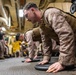 Military personnel in desert camouflage uniforms are performing push-ups on weight plates in what appears to be a ship's hangar bay or military vehicle bay. The foreground subject is wearing a plate carrier/tactical vest while doing push-ups with hands placed on circular weight plates. Additional service members are visible in the background, some observing and others participating in the physical training exercise.