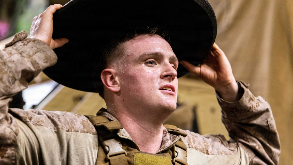 A young male service member in desert camouflage (MARPAT pattern) uniform and tactical vest is adjusting a dark wide-brimmed hat onto his head. He appears to be in a field or staging environment, looking upward with his mouth slightly open. The image is a close-up portrait-style photograph with good lighting and sharp focus on the subject.
