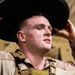 A young male service member in desert camouflage (MARPAT pattern) uniform and tactical vest is adjusting a dark wide-brimmed hat onto his head. He appears to be in a field or staging environment, looking upward with his mouth slightly open. The image is a close-up portrait-style photograph with good lighting and sharp focus on the subject.