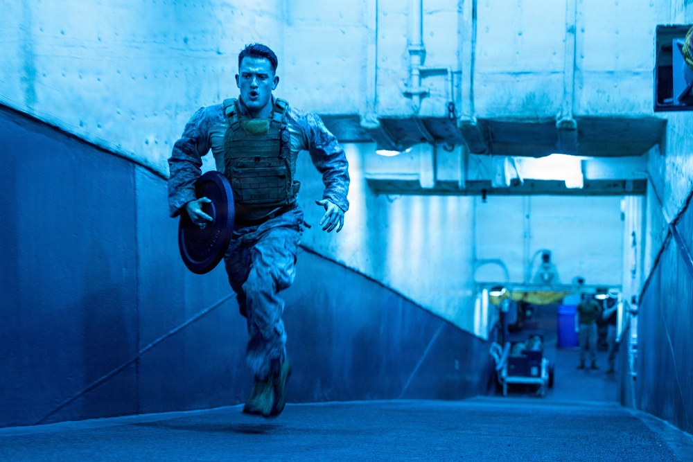 A military service member in camouflage uniform and tactical vest is running at full sprint through what appears to be an indoor corridor or tunnel, carrying a weight plate disc. The environment is bathed in a strong blue light, giving the scene a dramatic, high-intensity atmosphere. Additional personnel and equipment are visible in the background of the corridor.